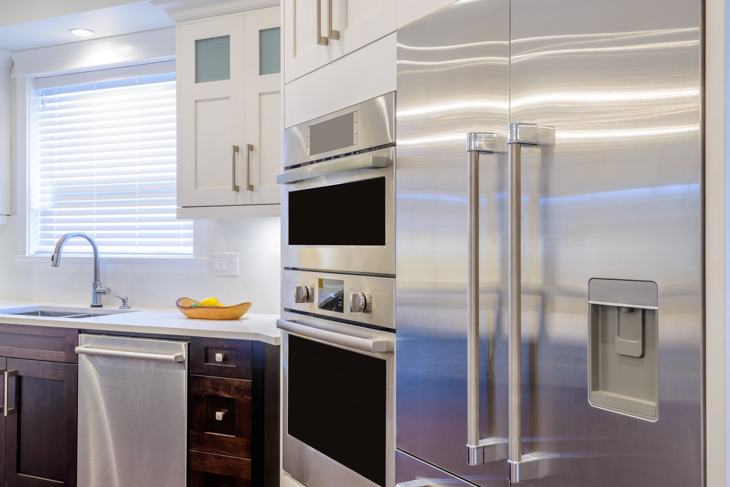 Modern Kitchen Interior With A Stainless Steel Refrigerator, Built-In Ovens, Dishwasher, And White Cabinets. A Sink Is Under A Window With Blinds, And A Fruit Bowl Is On The Countertop.