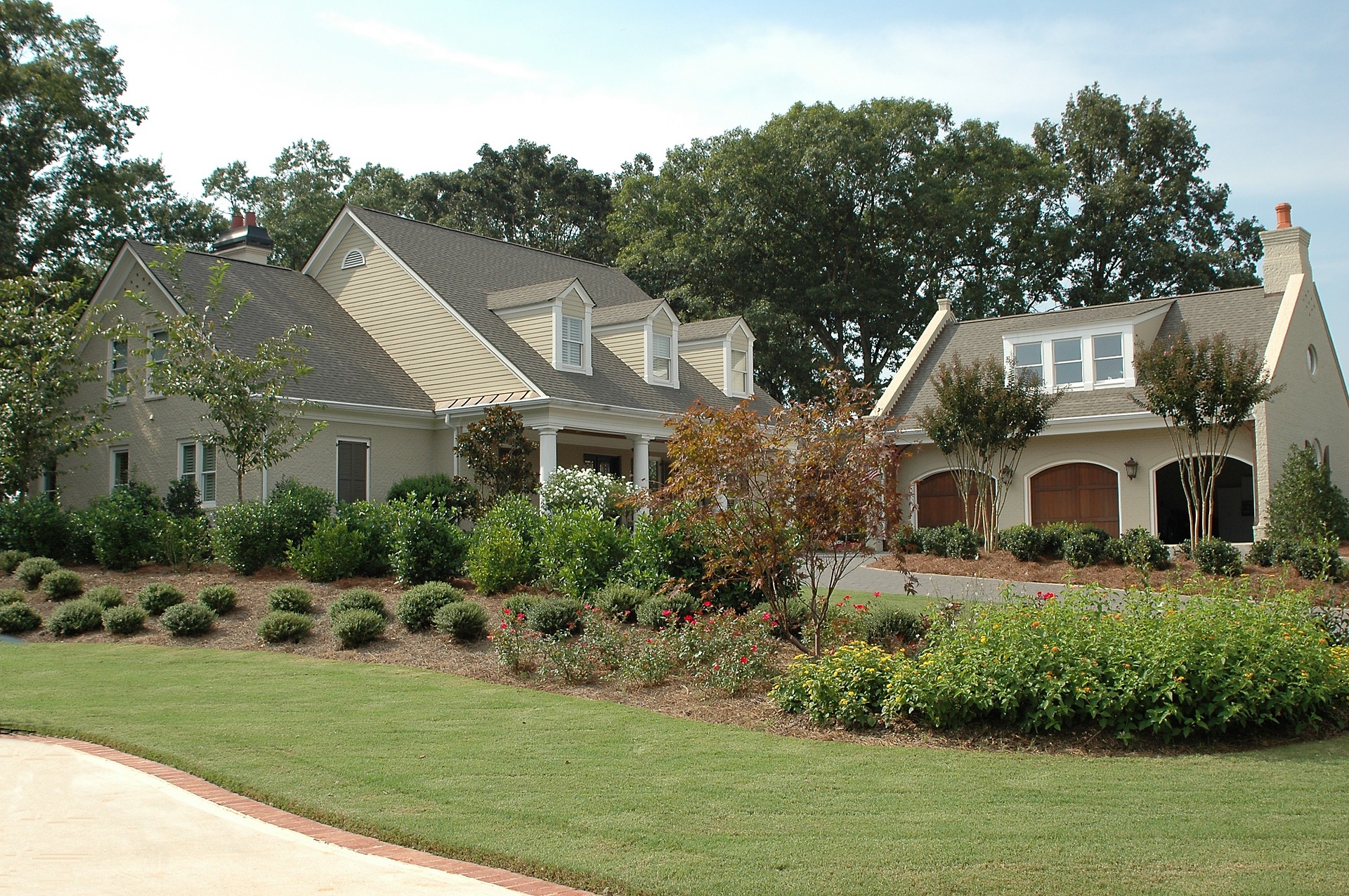 A Beige Two-Story House With A Sloped Roof, Surrounded By Manicured Lawn And Shrubs, With A Two-Car Garage Attached On The Right Side.