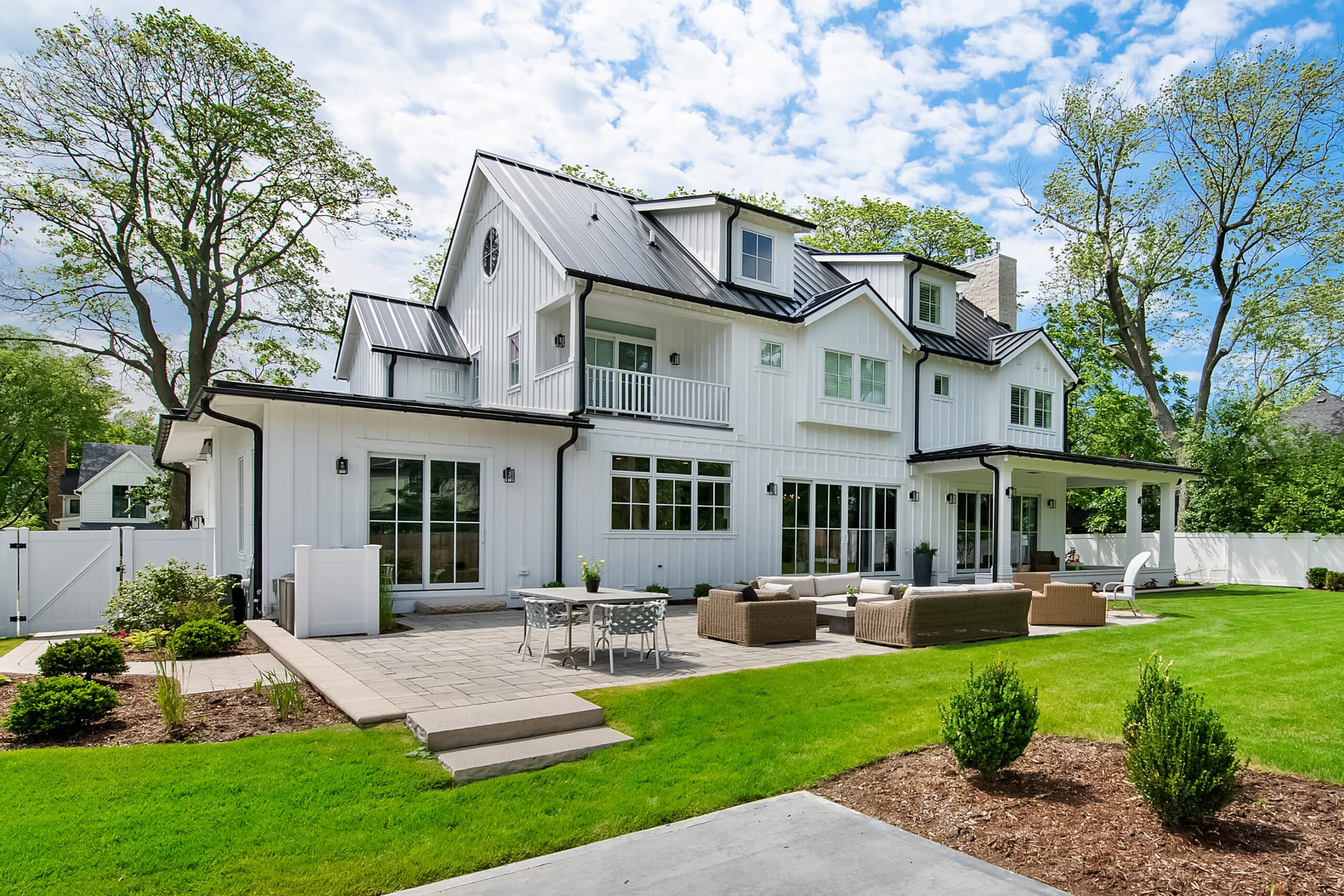 A Large White Two-Story House With Black Roof Accents, Surrounded By Trees. The Backyard Features Outdoor Seating And A Paved Patio Area With A Table And Chairs.
