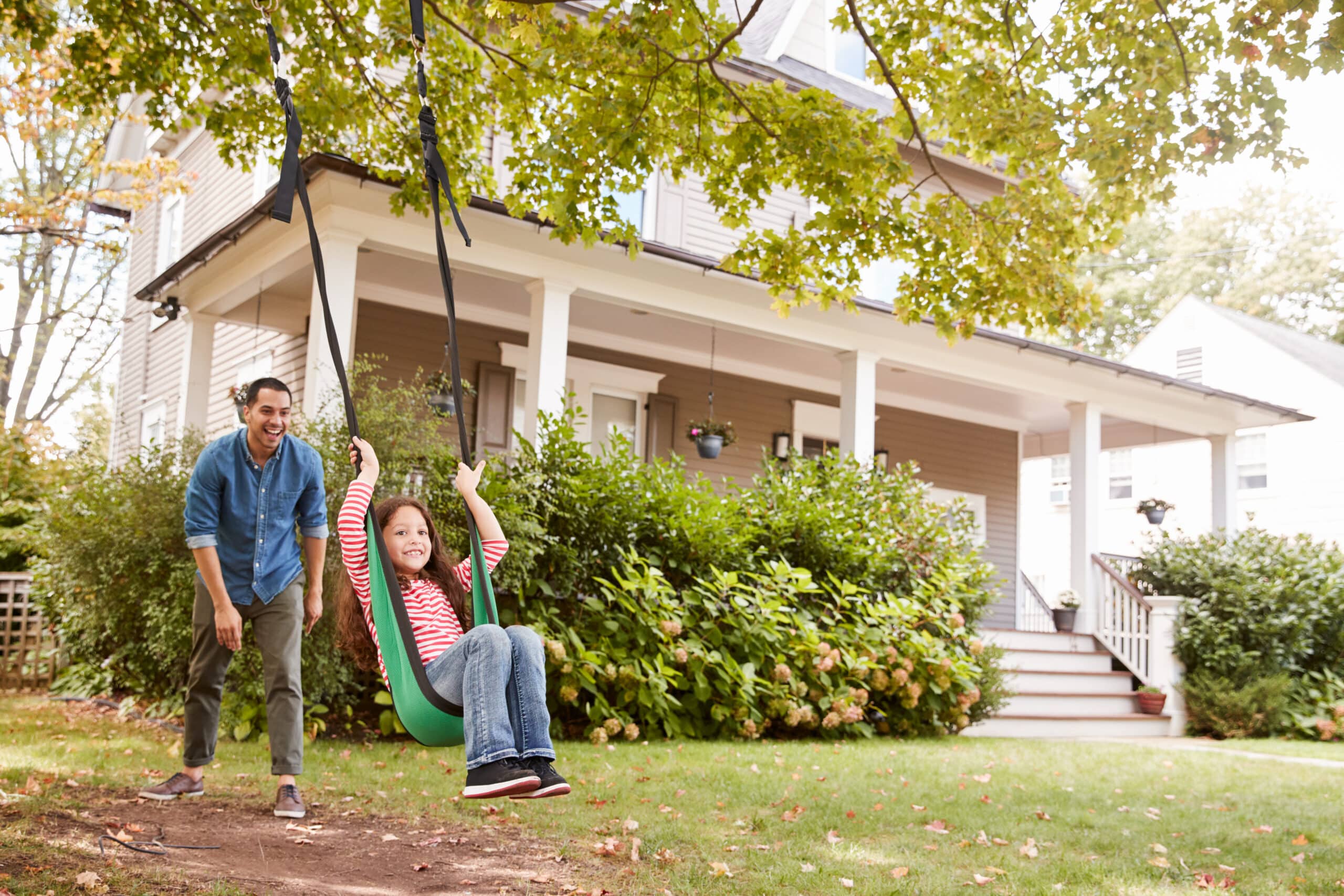 A Child In A Red And White Striped Shirt Is Swinging On A Tree Swing In Front Of A House. An Adult Is Pushing The Swing.