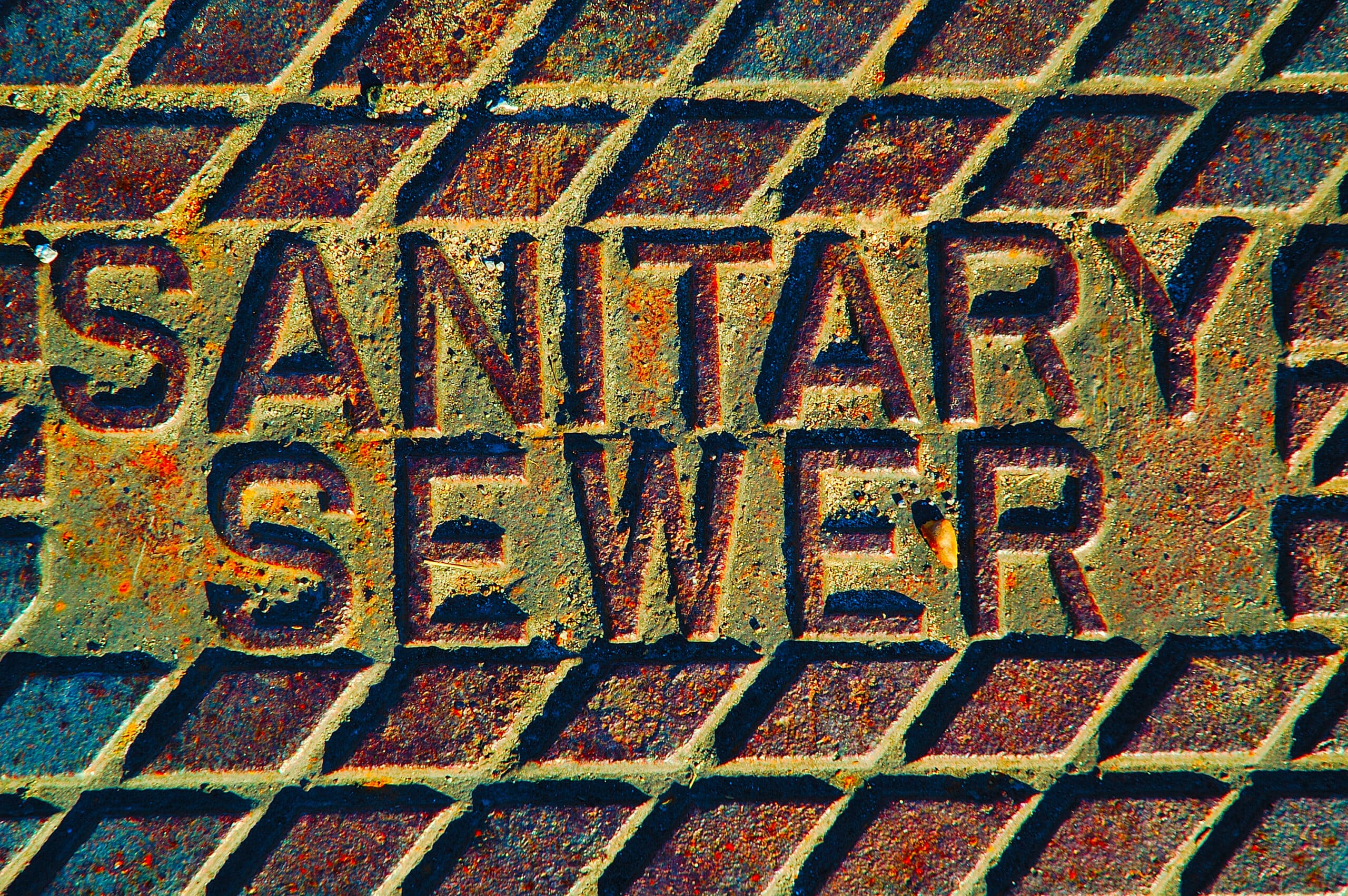 Close-Up Of A Metal Manhole Cover With The Words &Quot;Sanitary Sewer&Quot; Embossed On It, Featuring A Grid Pattern And Signs Of Rust.
