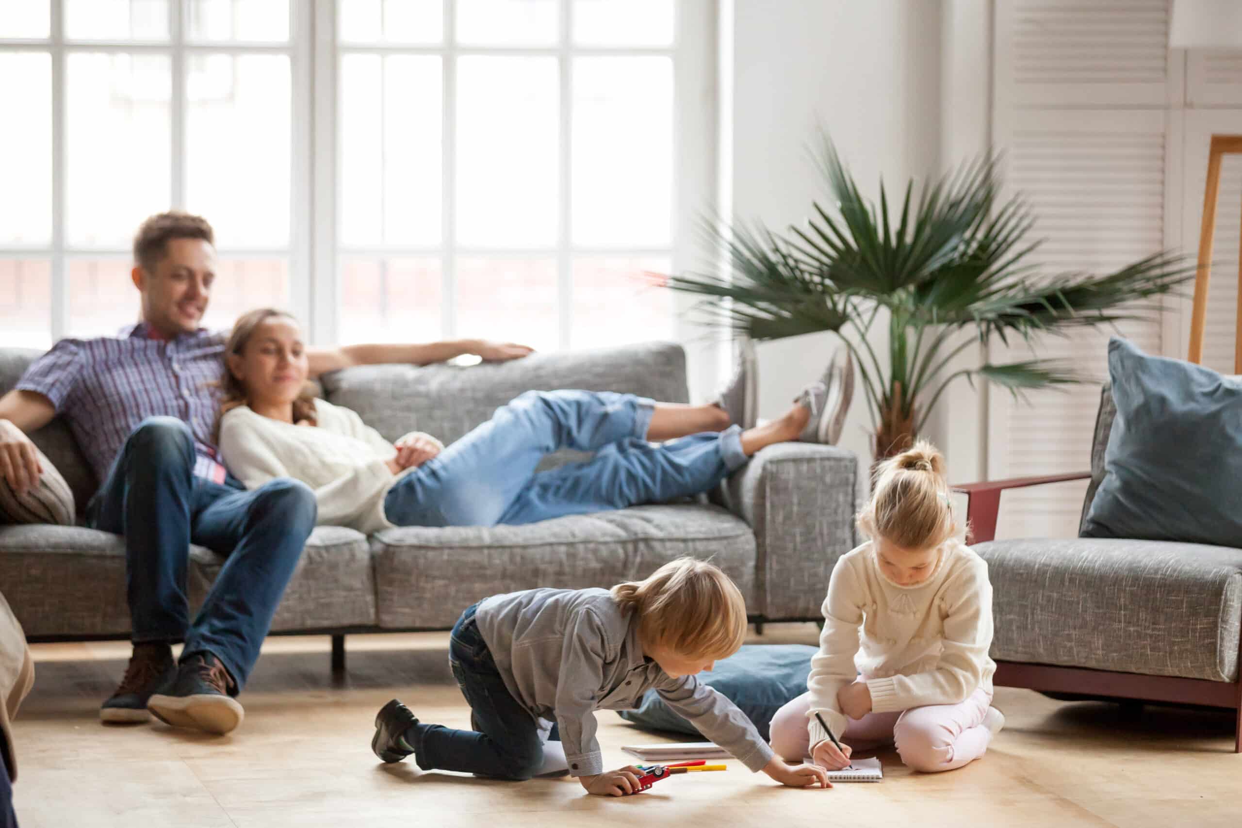 Two Children Draw On The Floor While A Man And Woman Relax On A Sofa In A Bright Room With Large Windows And A Potted Plant.