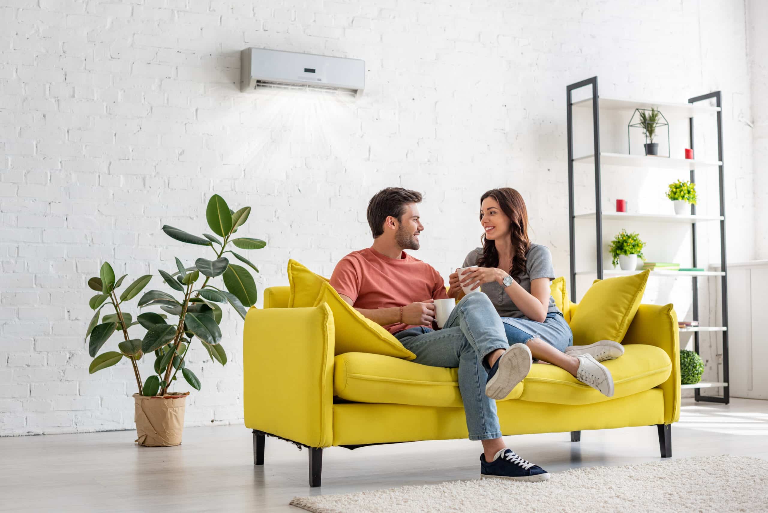 A Man And Woman Sit Talking On A Yellow Couch In A Bright Room With Plants, A Shelf, And An Air Conditioner Above Them.