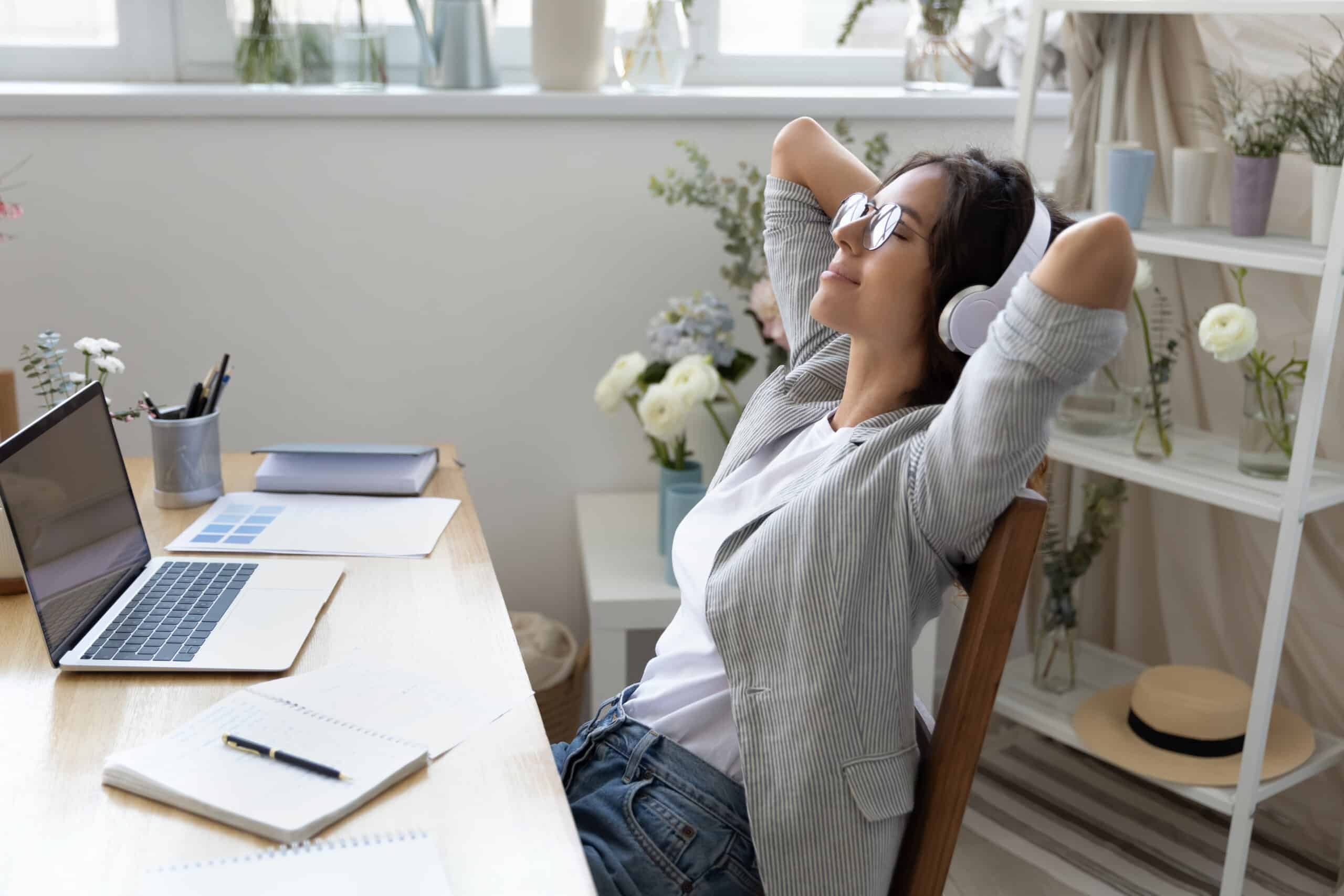 A Person Wearing Glasses And Headphones Leans Back In A Chair At A Desk With A Laptop, Notebooks, And Flowers, Appearing Relaxed.