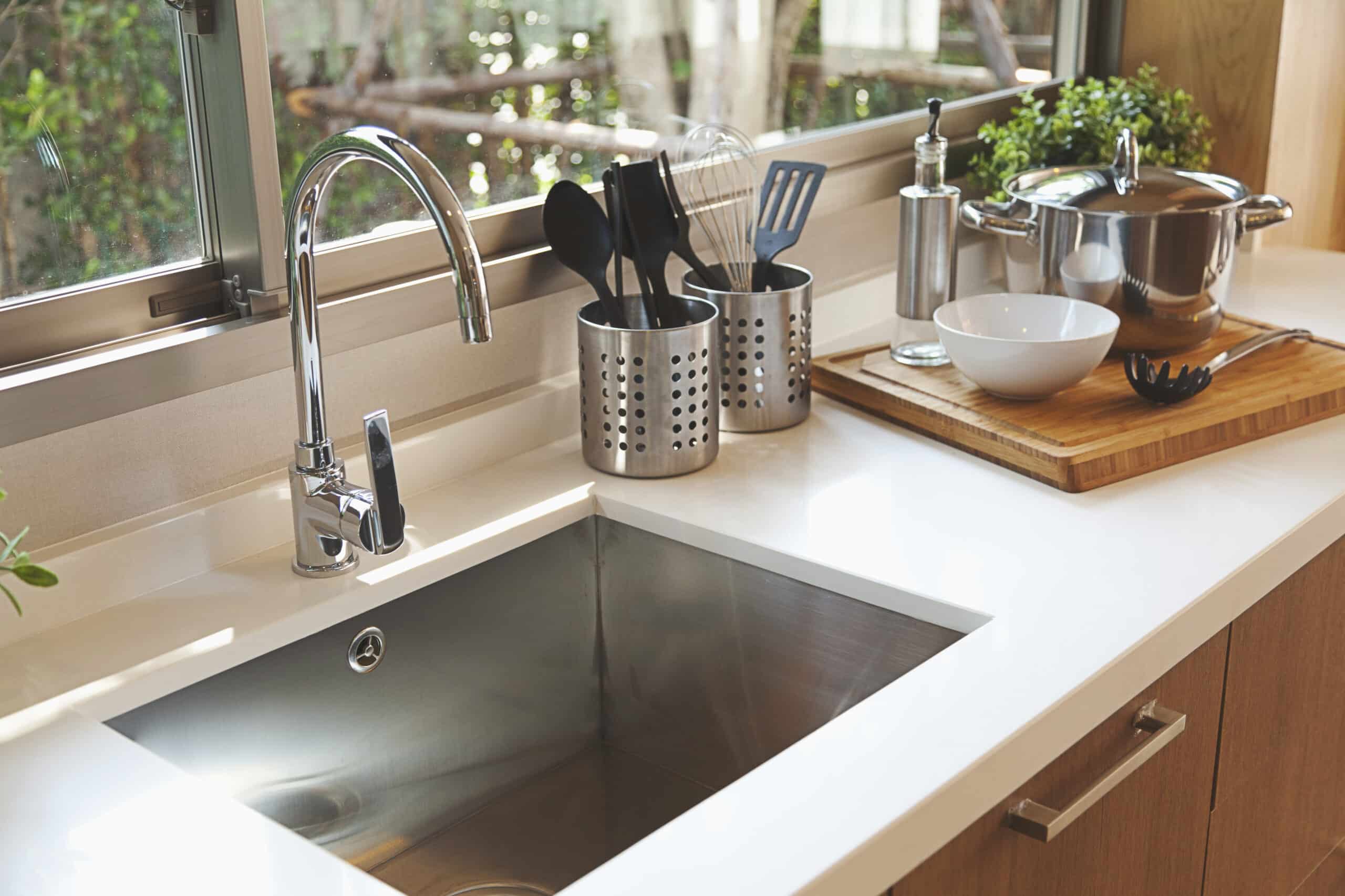 Modern Kitchen Sink With A Chrome Faucet. Nearby Are Containers Holding Utensils, A Chopping Board With A Pot And White Bowl, And Small Plants. Large Window In The Background.